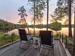 Une terrasse en bois avec deux chaises et une table, donnant sur un lac paisible au coucher du soleil.