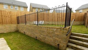 Un mur de soutènement en pierre, agrémenté d’une clôture métallique et d’un escalier menant à une zone herbeuse.