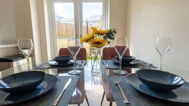 Une table à manger dressée pour quatre personnes, avec des assiettes noires, des couverts en argent et des verres à vin, et un vase rempli de tournesols placé au centre.