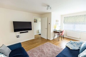 Un salón con un televisor de pantalla plana, un sofá azul, un tapete rosa y una ventana con persianas.