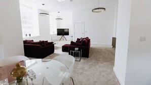 Ein modernes Wohnzimmer mit einer weißen Sofagarnitur, einem Glastisch und einem Fernseher auf einem Stativ.