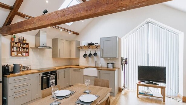 A modern kitchen with wooden beams, a dining table set for two, and a television in a corner. - Converted Barn near Lake District National Park (Carlisle)