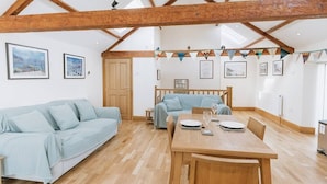 A spacious loft with exposed wooden beams, a dining area with a wooden table and chairs, a light blue sofa, and a loft bed with a view. - Converted Barn near Lake District National Park (Carlisle)