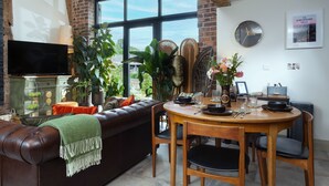 Una sala de estar acogedora con un sofá de cuero marrón, una mesa de comedor de madera con sillas, un televisor montado en la pared y ventanas amplias con vistas a zonas verdes.