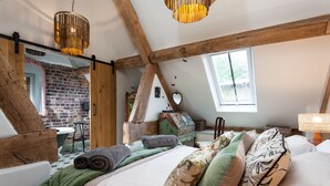 Un dormitorio con techo inclinado, vigas de madera a la vista, pared de ladrillo, una ventana y una cama con muchas almohadas y una manta verde.