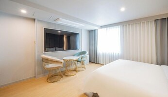 Habitación de hotel moderna con una cama grande, un área de estar con dos sillas y una mesita, un televisor montado en la pared y un ventanal con cortinas.