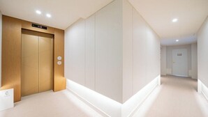 A modern hallway with a wooden elevator and a white wall with a vertical panel. - Denbastar Hotel-Kyungnam University Branch (Changwon)