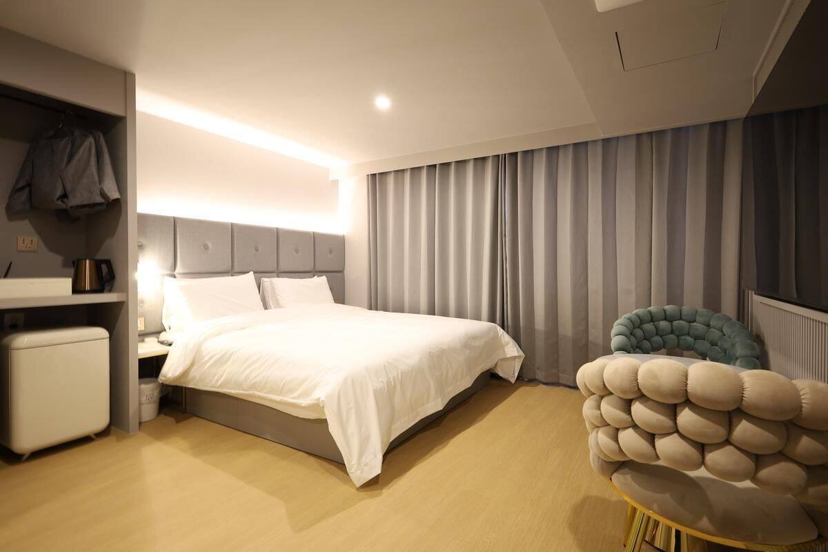 A modern hotel room with a large bed, a bedside table with a lamp, a chair, and a unique wall-mounted cushion.