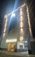 Exterior del hotel Den Basta con señalización iluminada y una 'P' destacada para estacionamiento.
