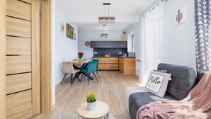 A modern living room with a grey sofa, a small round table with a plant, and a "Hello" pillow. In the background, there is a kitchen with a black countertop and wooden cabinets.