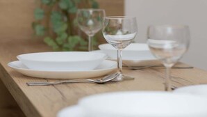 Ein Holztisch mit weißen Tellern und Weingläsern, vor einer Holzkopfteil und einer Grünpflanze mit rosafarbenen Blüten.