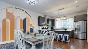 A modern kitchen with a central island, stainless steel appliances, and a dining area with white chairs and a patterned rug. - Modern Townhome w/ Game Room + 10min to Downtown! (Nashville)