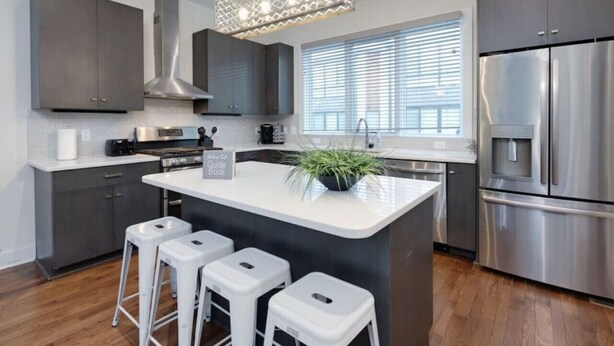 A modern kitchen with grey cabinets, a stainless steel refrigerator, a central island with stools, and a hanging light fixture. - Modern Townhome w/ Game Room + 10min to Downtown! (Nashville)