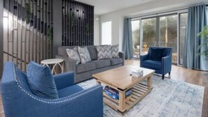 A modern living room with a grey sofa, blue armchairs, a wooden coffee table, and a large window with curtains. - Modern Townhome w/ Game Room + 10min to Downtown! (Nashville)