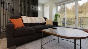 En moderne stue med en sort sofa, et træbord og et stort vindue med udsigt til træer.