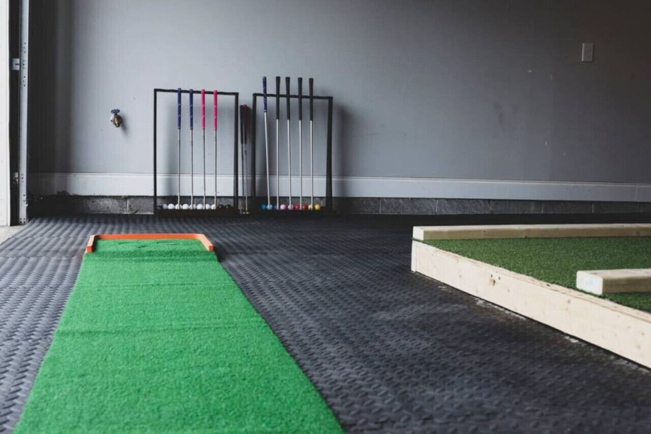 A room with a green carpet, a wooden playpen, and a wall-mounted rack with pool cues.