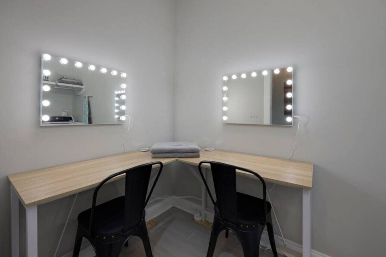Een kamer met een houten bureau, twee zwarte stoelen en twee spiegels met verlichting, bevestigd aan de muur.