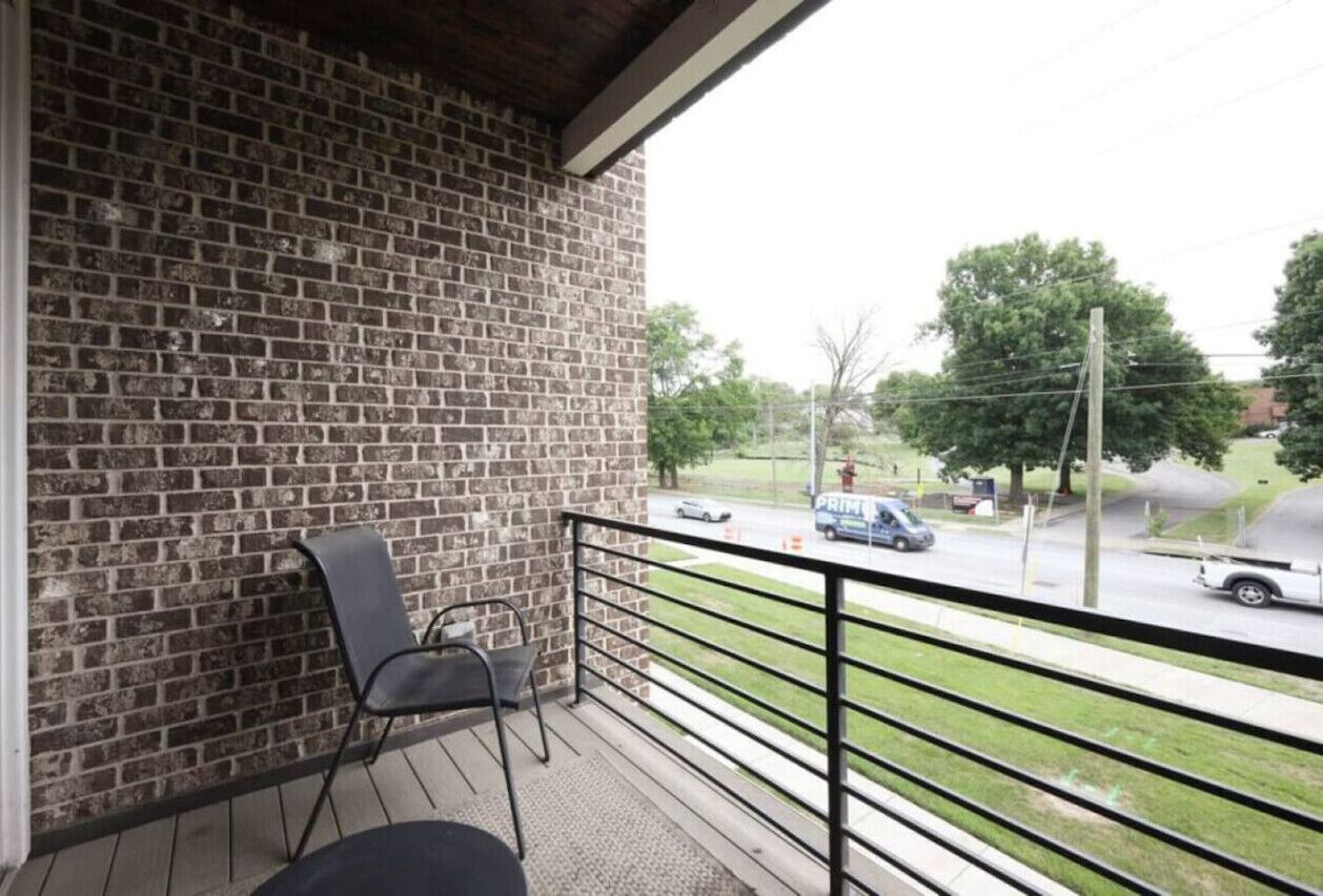 Un balcon avec un mur en briques, une chaise et une vue sur une rue bordée d’arbres et où circulent des voitures.