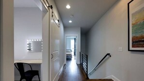 A hallway with a wooden floor, a black chair, a desk with a mirror, and a staircase with a handrail.