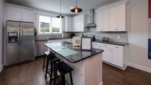 A modern kitchen with white cabinets, a stainless steel refrigerator, a central island with stools, pendant lighting, and a window providing natural light.