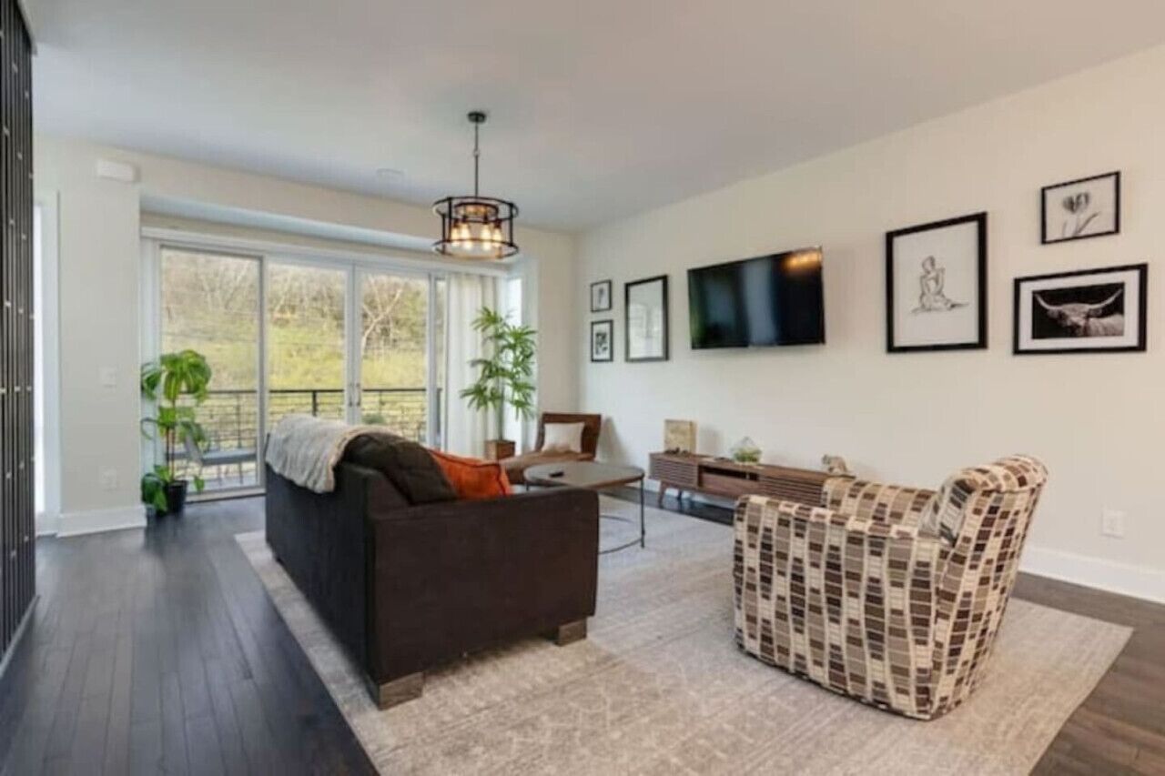 A spacious living room with a dark brown sofa, a patterned armchair, and a coffee table. There is a large window with a view of greenery outside.