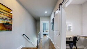 A hallway with wooden flooring, a staircase, a bed in the background, and a desk with a chair.