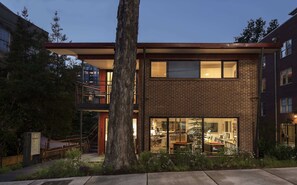 A modern two-story building with a brick facade, large windows, and a visible interior with furniture.