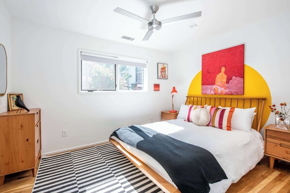 A bedroom with a bed, a ceiling fan, a window, a wooden dresser, and a painting on the wall.