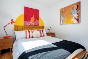 A bedroom with a bed, a nightstand, a red lamp, and two paintings on the wall.