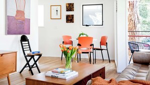 Un salon moderne avec une table basse en bois, un canapé et un coin repas avec des chaises orange. Un tableau orne le mur et une fenêtre offre une vue sur les arbres.