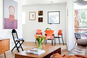 A modern living room with a wooden coffee table, a sofa, and a dining area with orange chairs. There is a painting on the wall and a window with a view of trees.