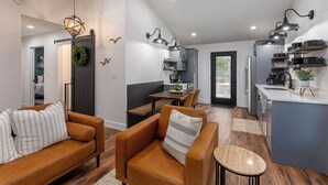 A modern living room with a brown leather sofa and a small round table, a kitchen with grey cabinets and a white countertop, and a dining area with a wooden table and chairs.