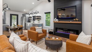 A modern living room with a fireplace, a TV mounted on the wall, and a dining area with a kitchen in the background.