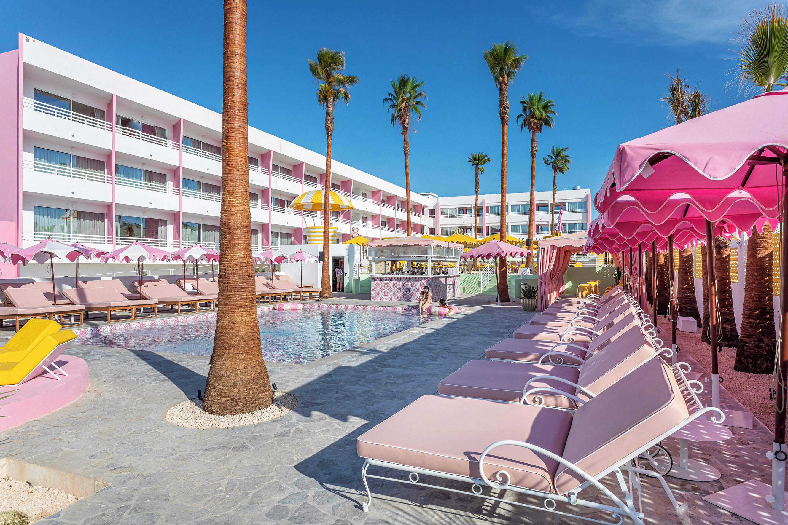 A pool area with lounge chairs and umbrellas, surrounded by palm trees and a multi-story building with balconies.