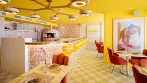 Een restaurant met een geel en wit kleurenschema, met een bar, verschillende zitmogelijkheden zoals barstoelen en tafels, en een moderne plafondlamp.