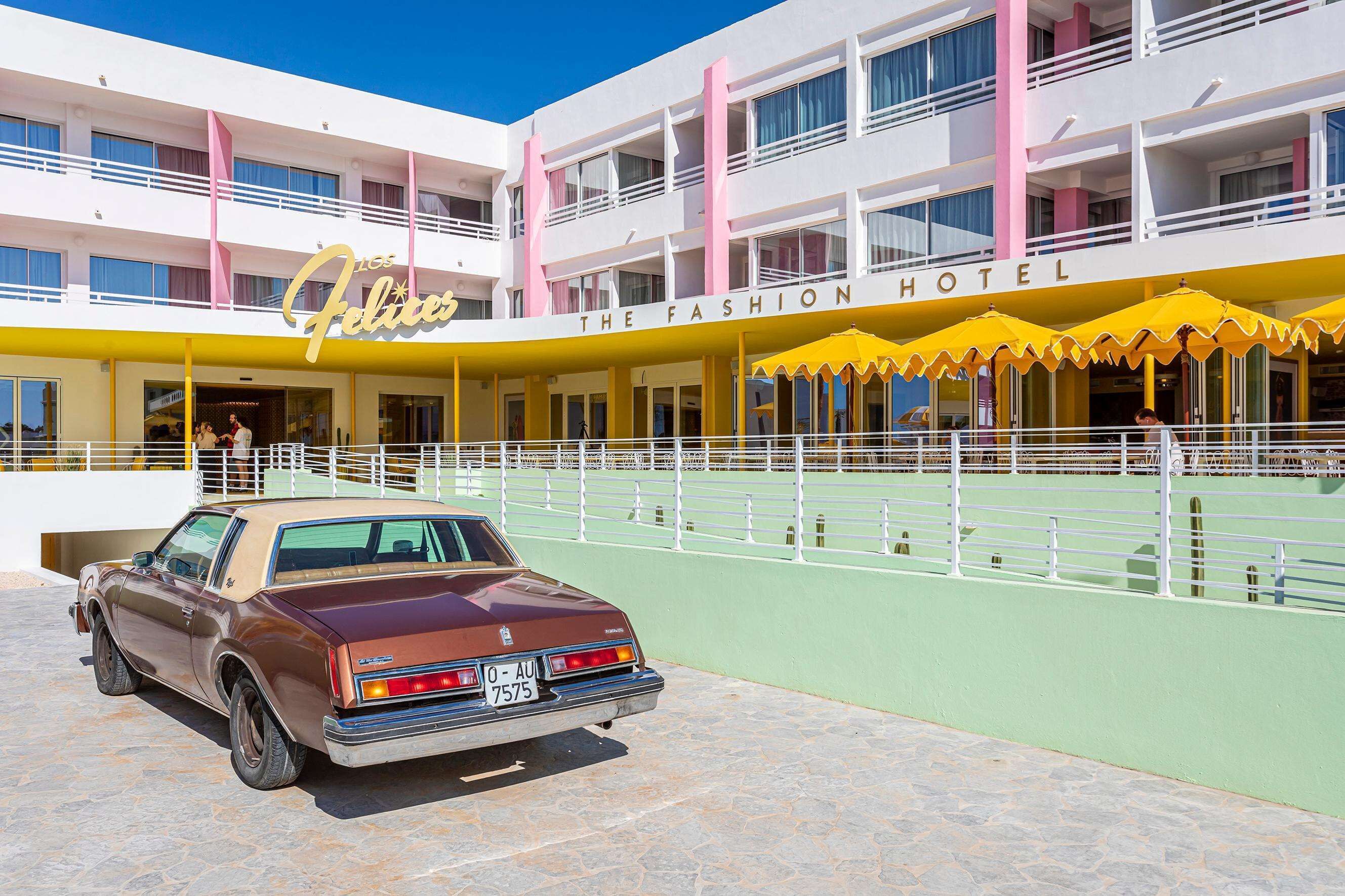 A hotel with a yellow and pink facade, a vintage car parked in front, and outdoor seating with yellow umbrellas.