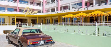 A hotel with a yellow and pink facade, a vintage car parked in front, and outdoor seating with yellow umbrellas.