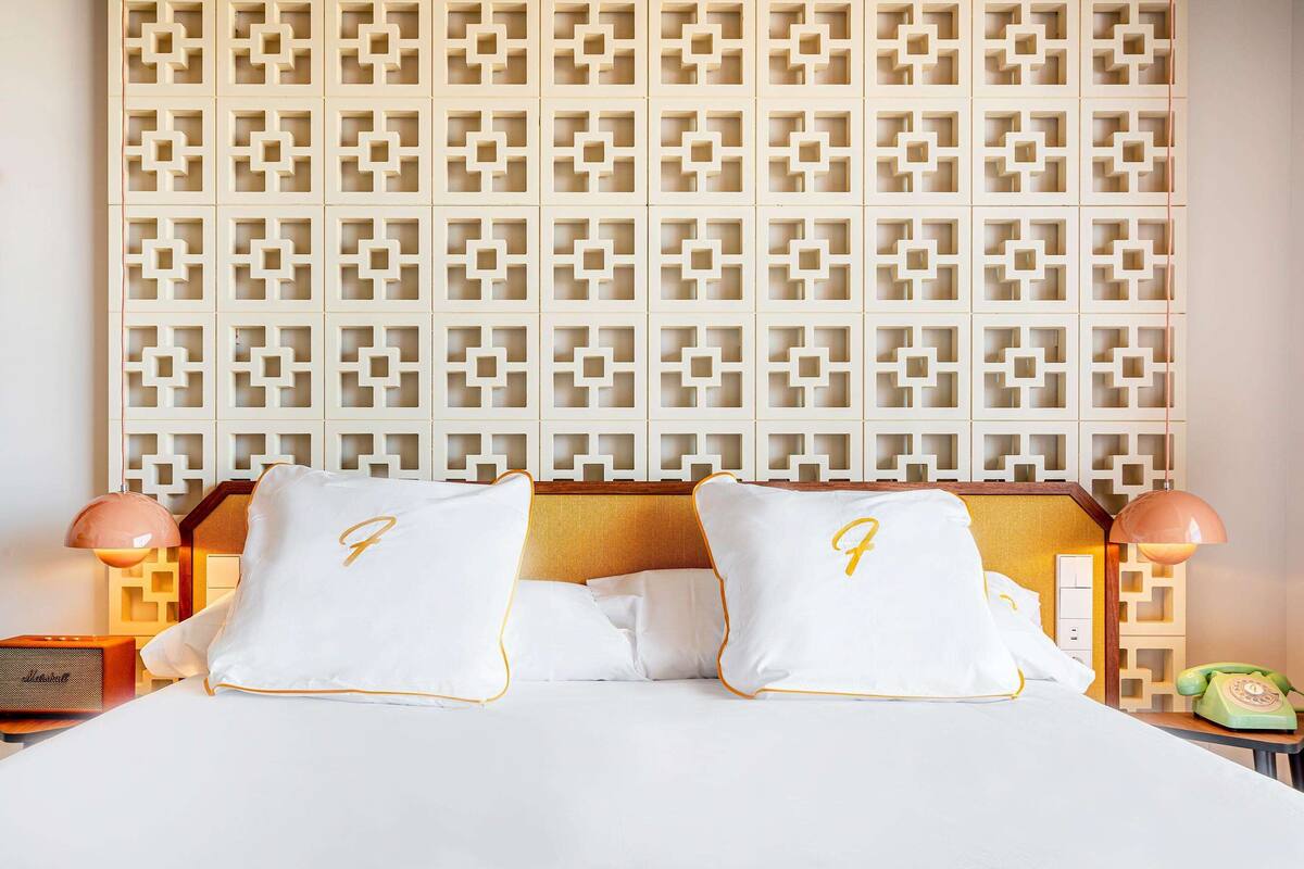 A hotel room with a bed, two pillows with a monogram, a nightstand with a lamp, and a telephone.