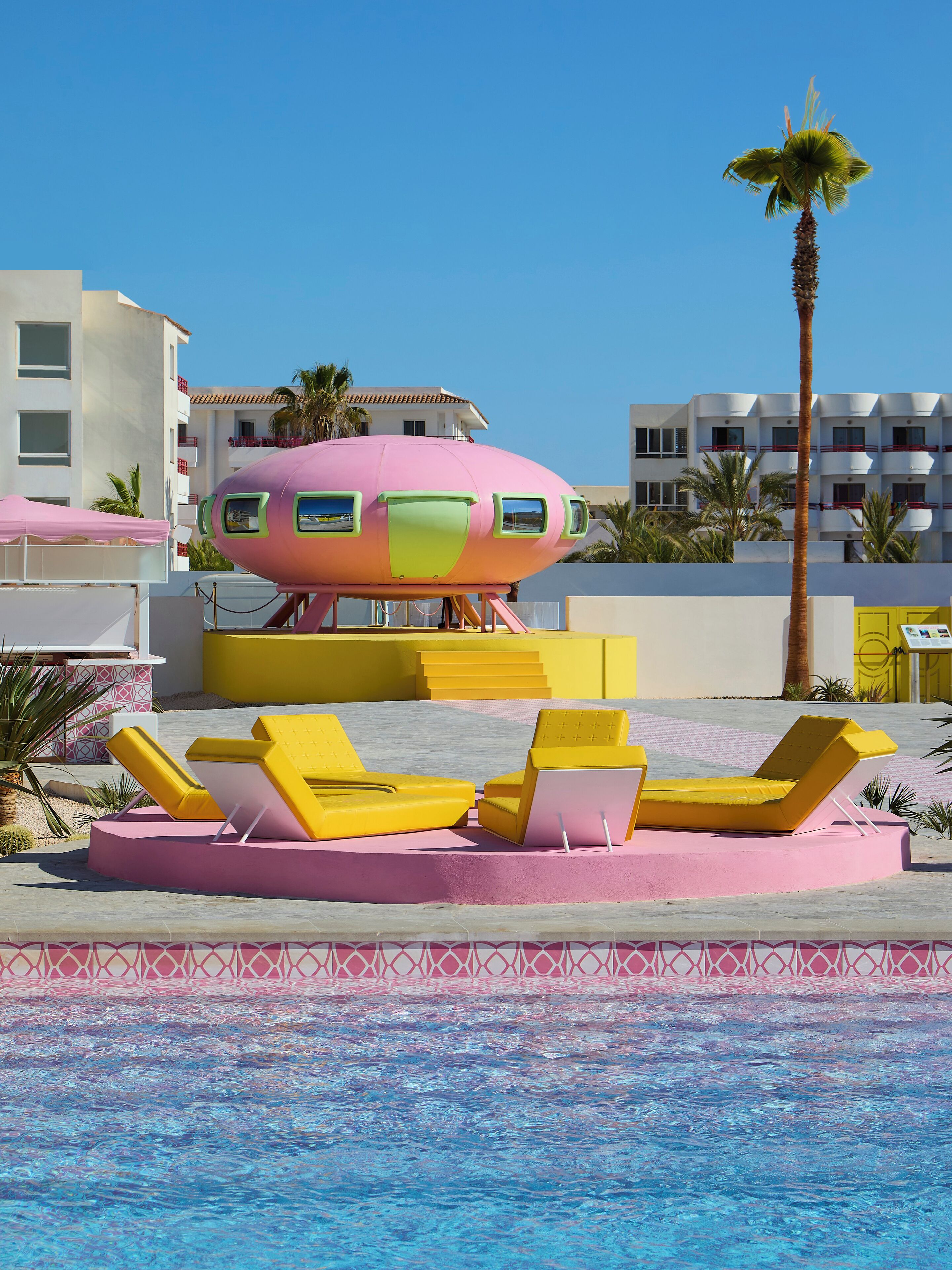 A pool area with yellow lounge chairs and a pink and green structure resembling a submarine.