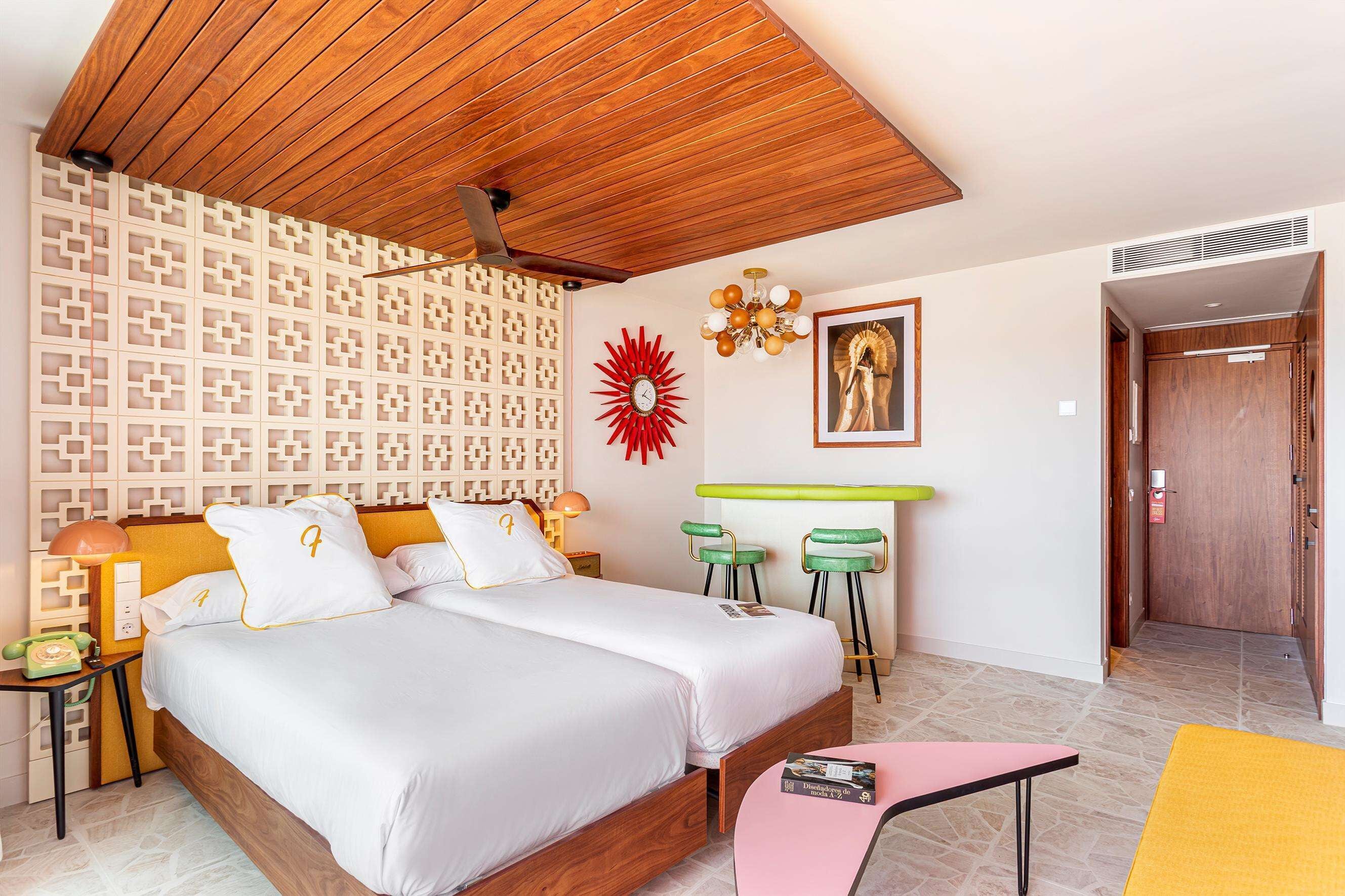Habitación de hotel con techo de madera, dos camas, pared con estampado, adorno de sol rojo, lámpara de araña, barra verde y una puerta que da a otra habitación.