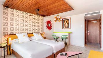 Habitación de hotel con techo de madera, dos camas, pared con estampado, adorno de sol rojo, lámpara de araña, barra verde y una puerta que da a otra habitación.