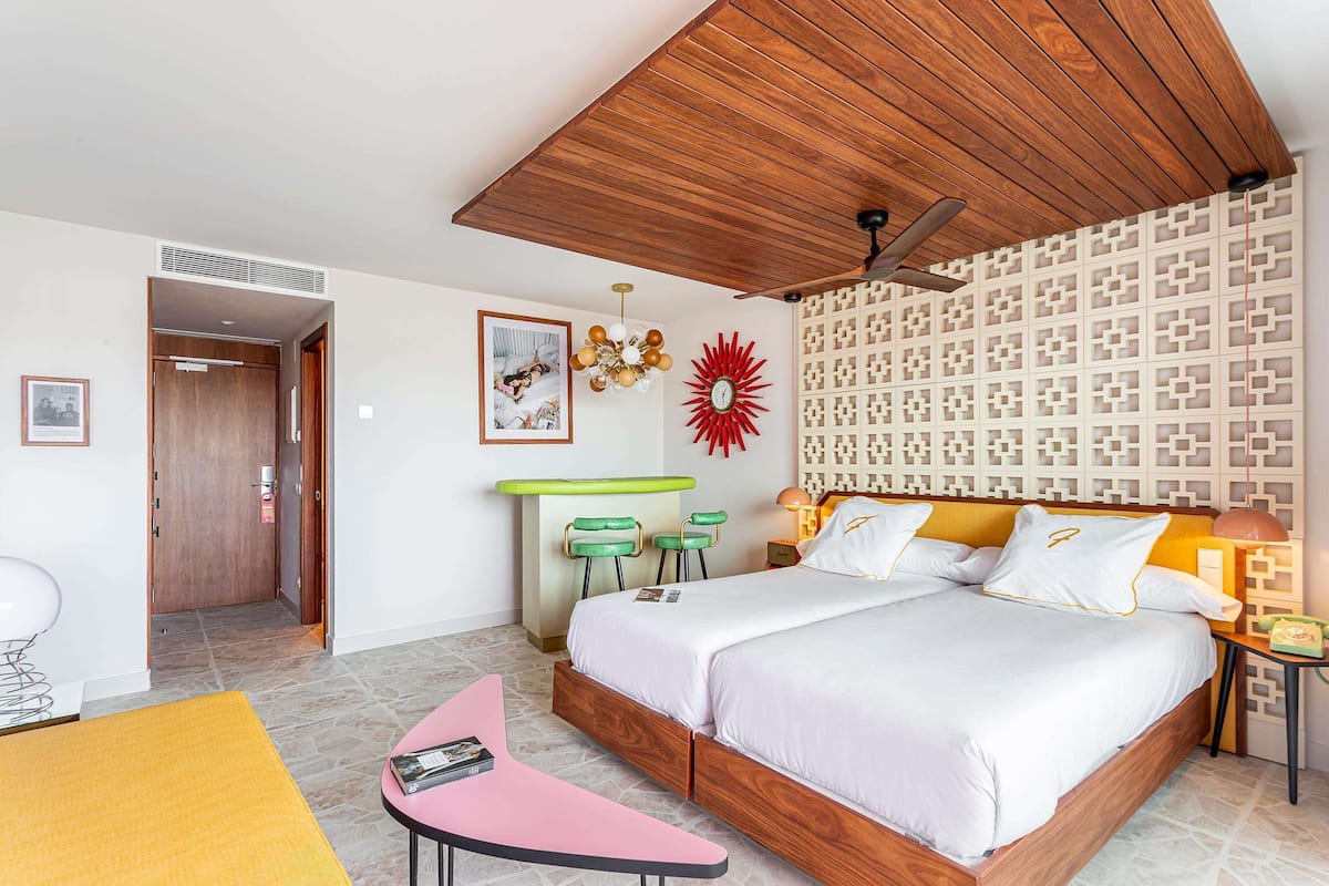 A modern hotel room with a wooden ceiling, a bed with white bedding, a decorative wall panel, and a ceiling fan.