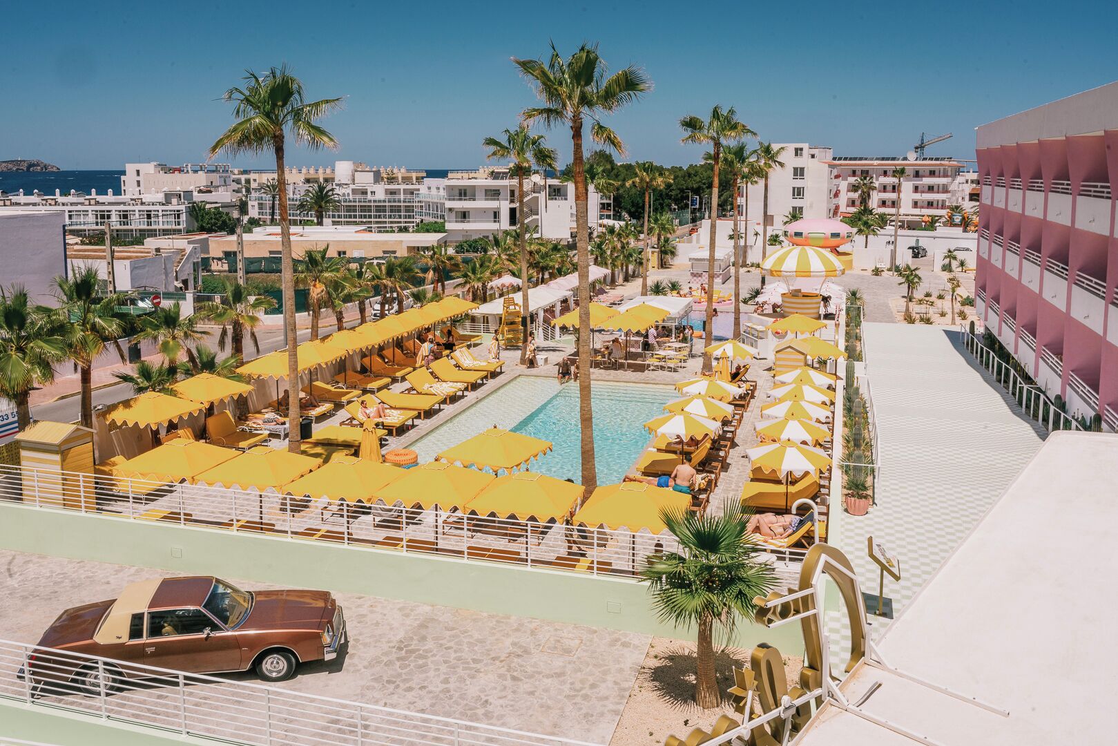 A resort with a swimming pool surrounded by sun loungers and umbrellas, a parking area, and a view of the sea and buildings in the background.