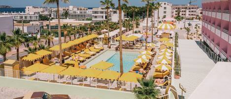 A resort with a swimming pool surrounded by sun loungers and umbrellas, a parking area, and a view of the sea and buildings in the background.