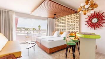 Habitación de hotel con una cama grande, un balcón con vistas, ventilador de techo, un reloj de pared decorativo y una zona de bar con taburetes.