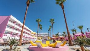 Een resort met een roze en witte gebouw, palmbomen en gele ligstoelen.