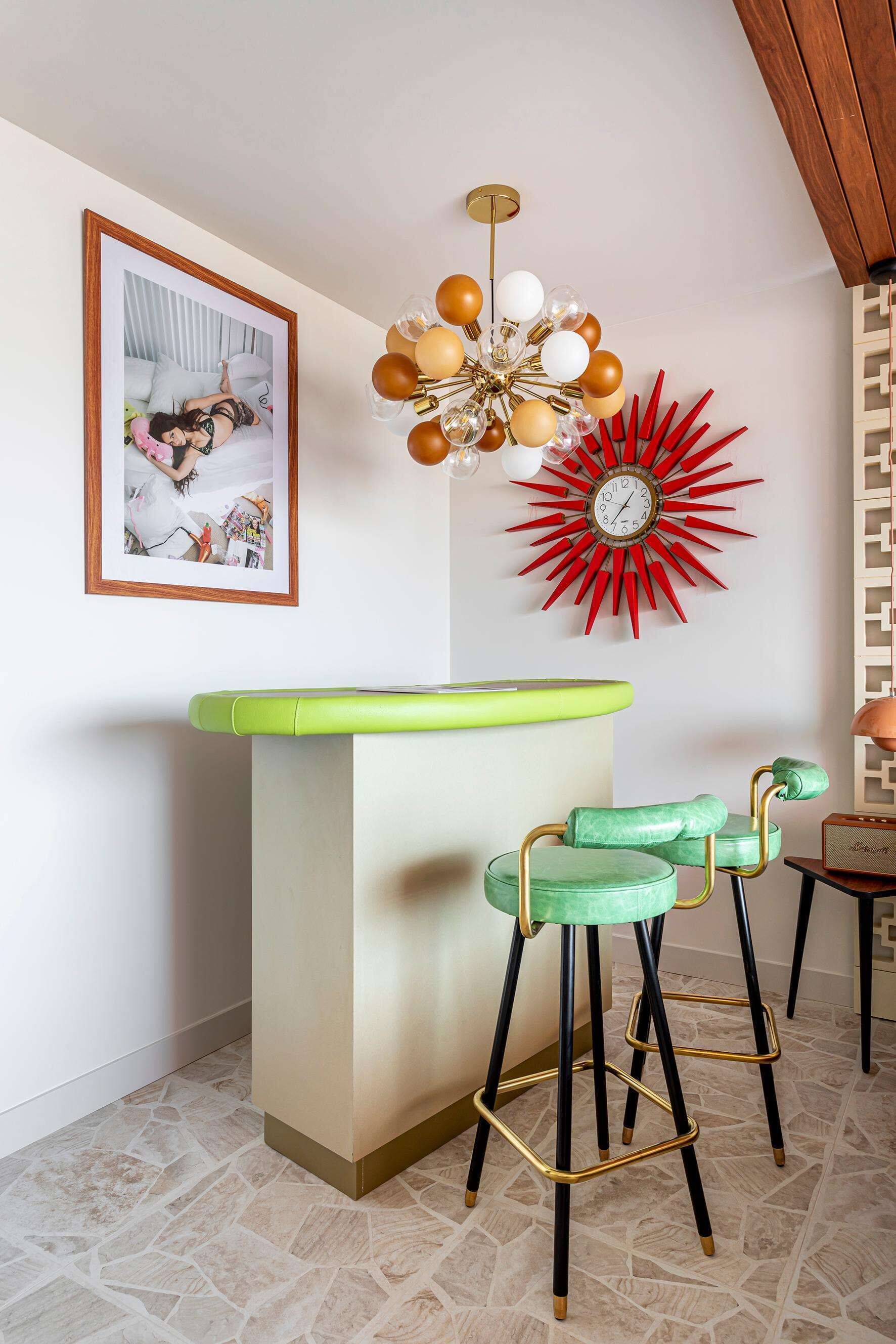 A bar area with a green countertop, two green bar stools with black and gold legs, a red sunburst clock, and a framed photo on the wall.