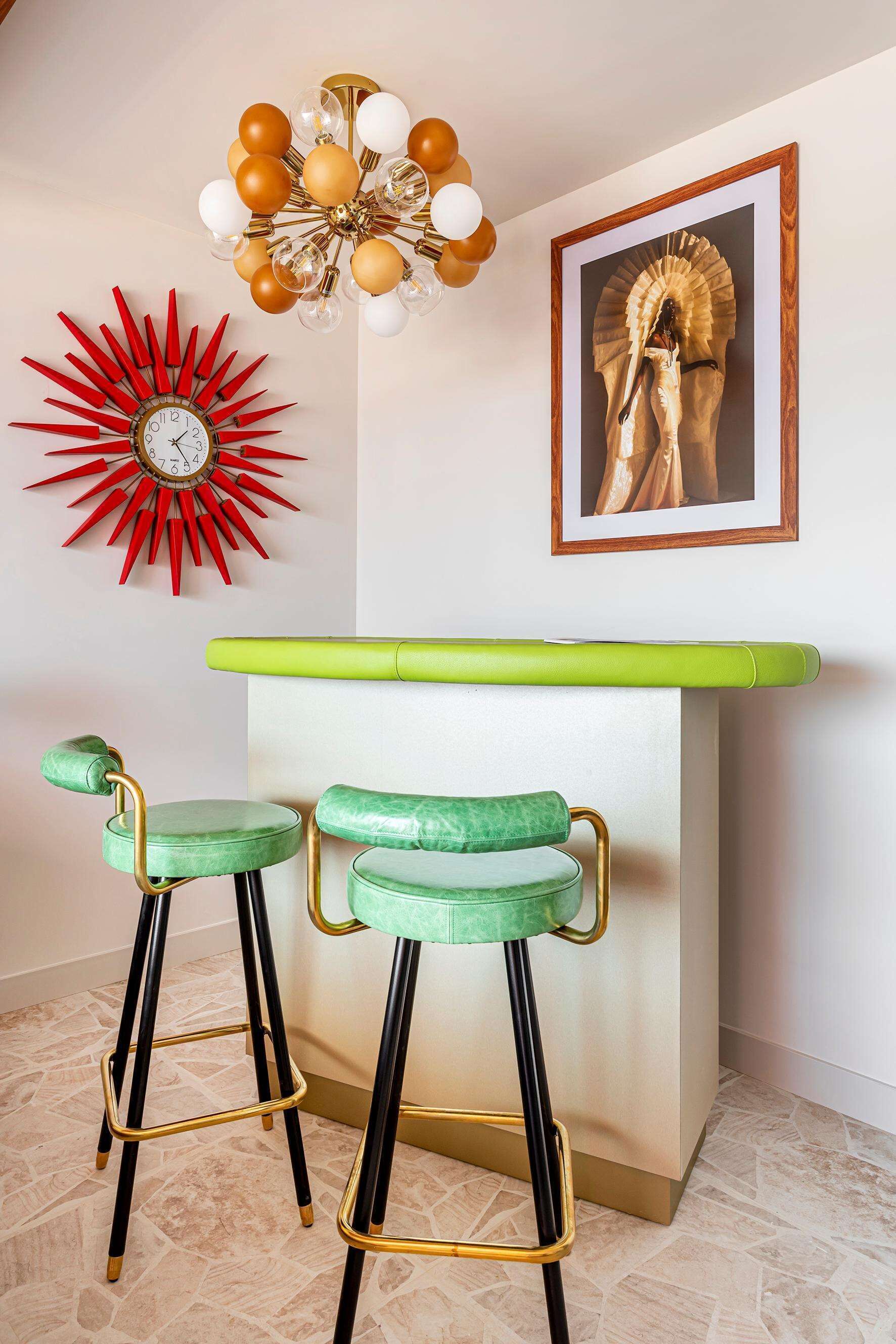 A bar area with two green bar stools, a red sunburst clock, a gold-framed chandelier, and a framed artwork on the wall.