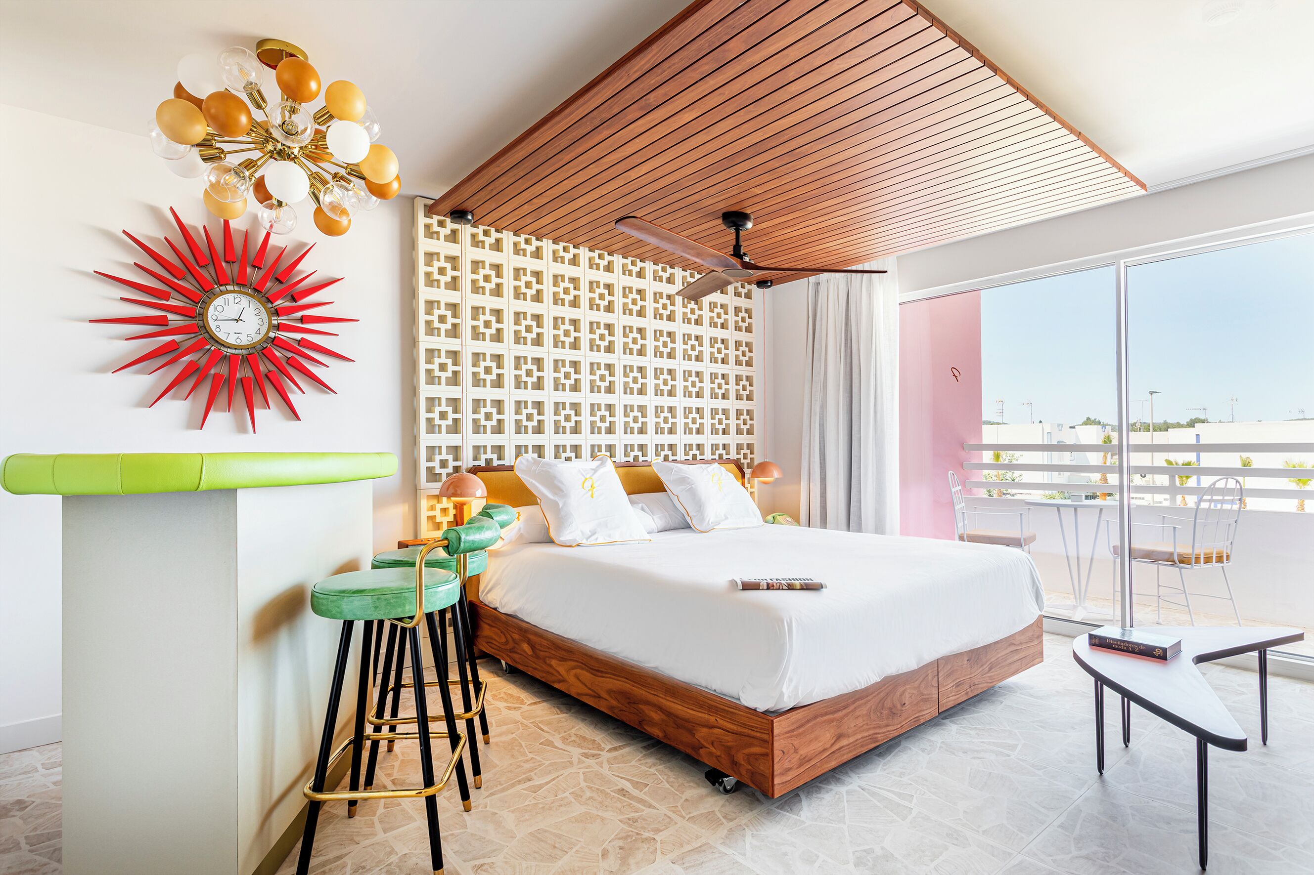 A modern hotel room with a wooden ceiling, a red sunburst clock, a green bar stool, a bed with white bedding, and a balcony with a view.