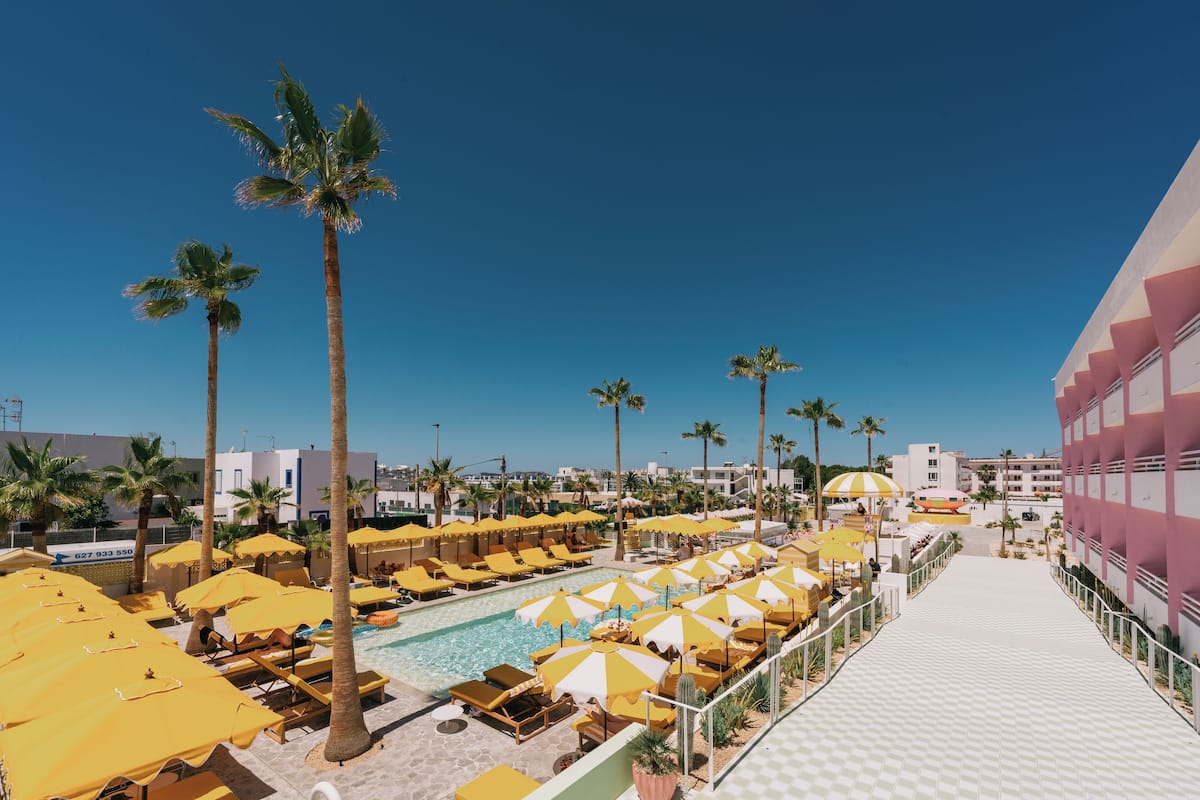 A pool area with yellow umbrellas and lounge chairs, surrounded by palm trees and a clear blue sky.